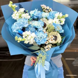 A stunning bouquet titled "Mystic Blue Charm," featuring elegant blue carnations, delicate matthiolas, and striking anemones, beautifully arranged and wrapped in eco-friendly packaging.